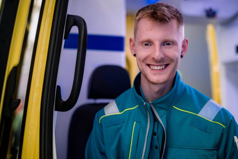 Leihkraft Keanu Will lächelt und berichtet über Zeitarbeit im Rettungsdienst. Er trägt eine blaue Jacke und die Uniform des Notdienstes