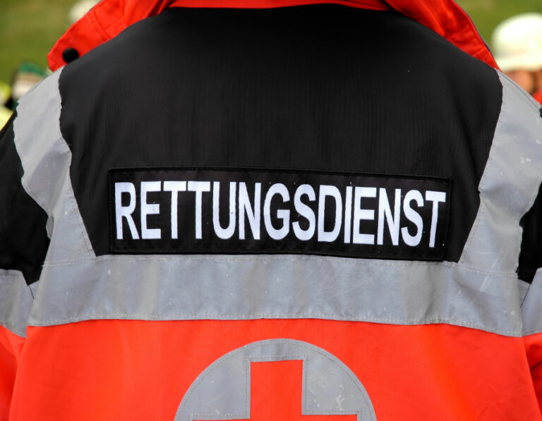 Die Rückseite einer Rettungsdienst-Jacke ist zu sehen. Auf der schwarz hinterlegten Fläche steht in weißer Schrift „RETTUNGSDIENST“. Die Jacke ist in den Farben Schwarz, Grau und Signalorange gestaltet und trägt am unteren Rand das Symbol eines roten Kreuzes.